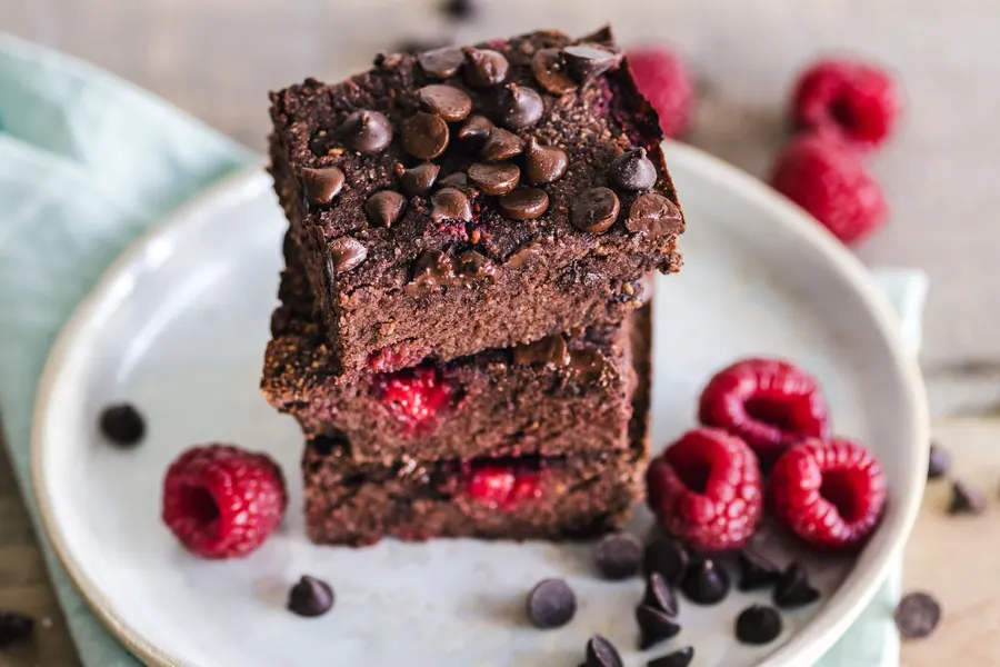A stack of fudgy chocolate brownie squares topped with chocolate chips, plated with fresh raspberries