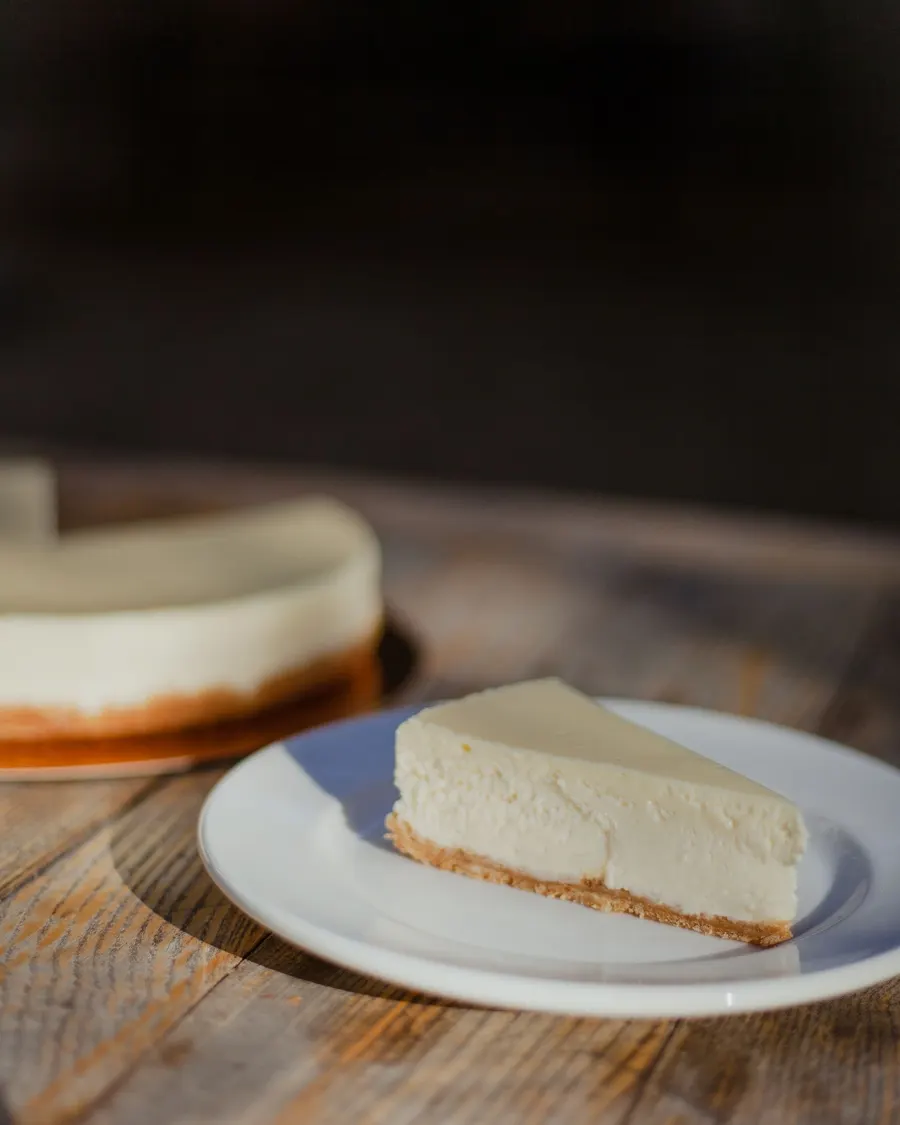 A plain slice of New York-style cheesecake on a white plate, the whole cake blurred in the background